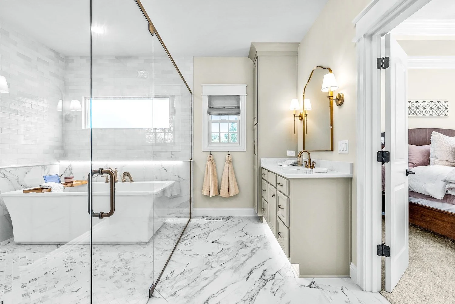 A luxurious bathroom with a glass-enclosed shower, white marble flooring, a bathtub, a small window, beige towels, a vanity with a mirror and two wall-mounted lamps, and a glimpse into a bedroom with a bed and pillows.