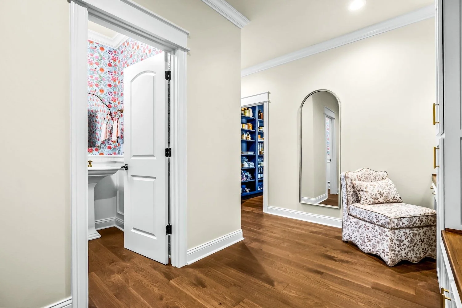 Interior view of a home hallway with a floral armchair, a tall mirror, and open doorways showing a bathroom with floral wallpaper and a blue bookshelf.