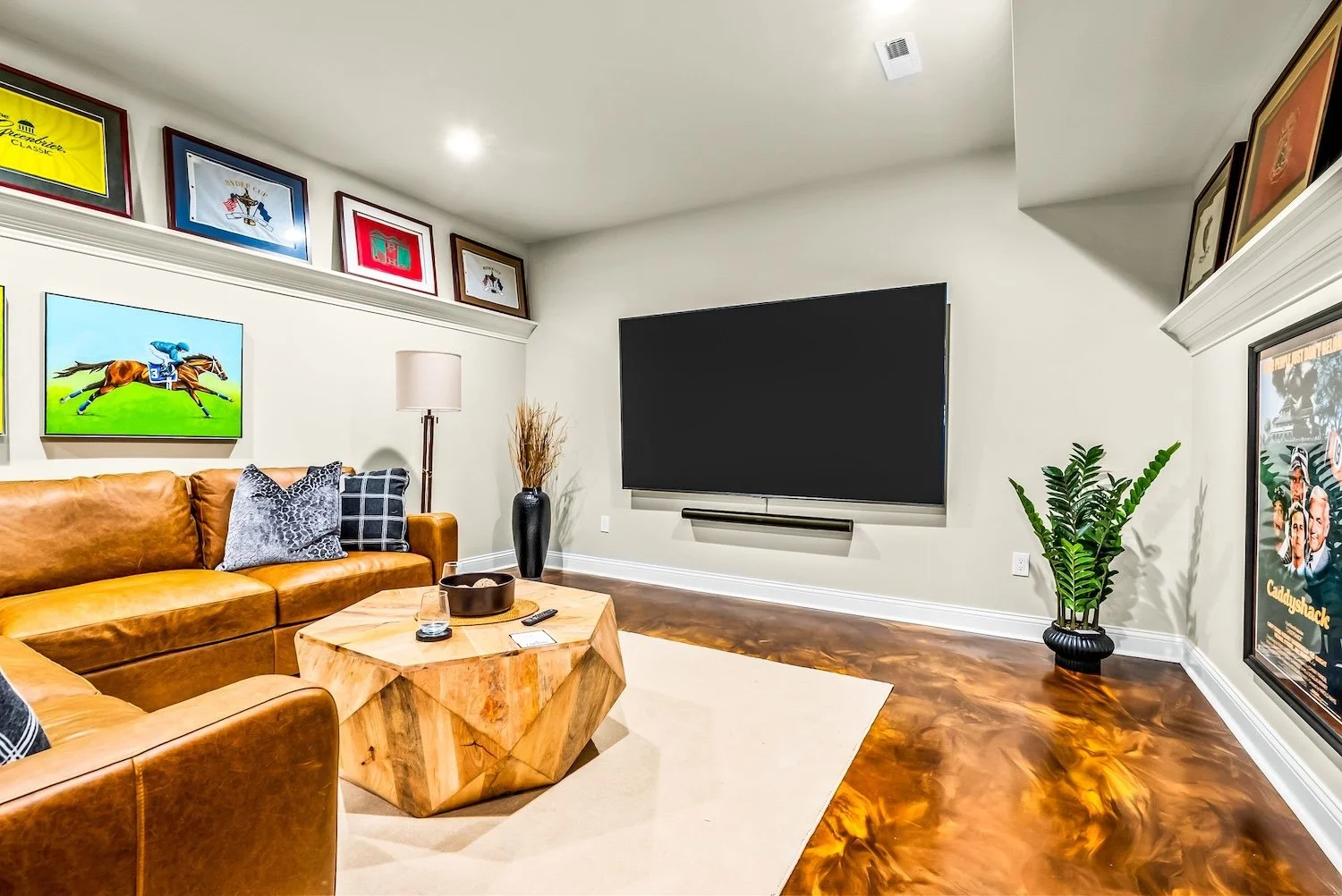 Living room with a leather sectional sofa, a large flat-screen TV on the wall, framed artwork and posters, a tall plant in a black pot, a wooden coffee table with a tray and remote controls, a beige rug, and decorative items including pillows, a lamp