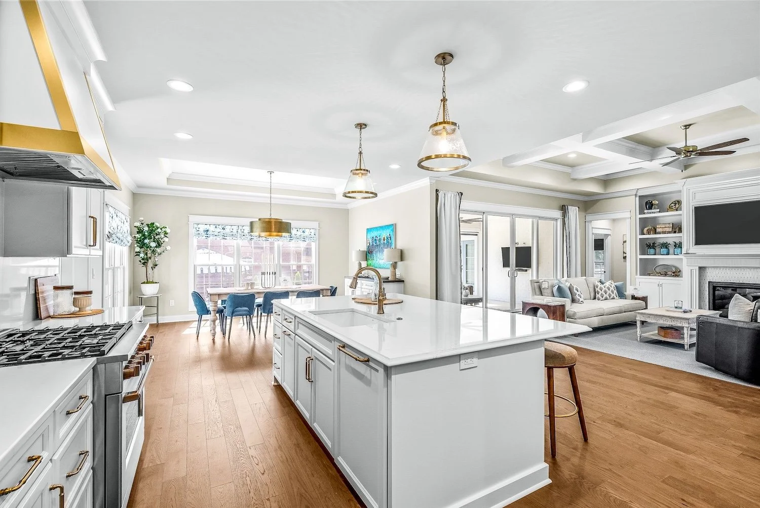 Open-concept kitchen, dining, and living room with white cabinets, gold accents, hardwood floors, and large windows letting in natural light.