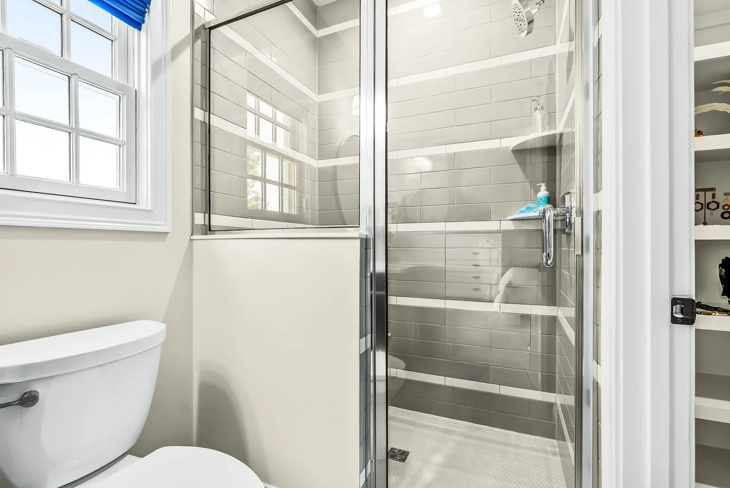 Bathroom with a toilet, a glass shower enclosure with gray tiles, a window with white trim, and a white shelf with toiletries.