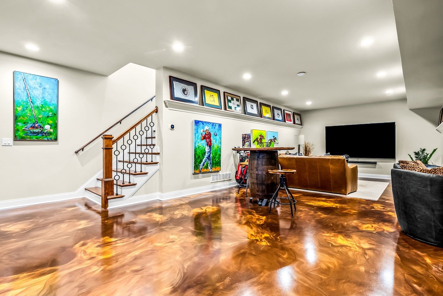 A spacious living room with polished, swirling wood flooring, featuring a staircase with wooden handrail and metal balusters, a large flat-screen TV mounted on the wall, a brown leather sofa, a black armchair with leopard print pillow, a small round 