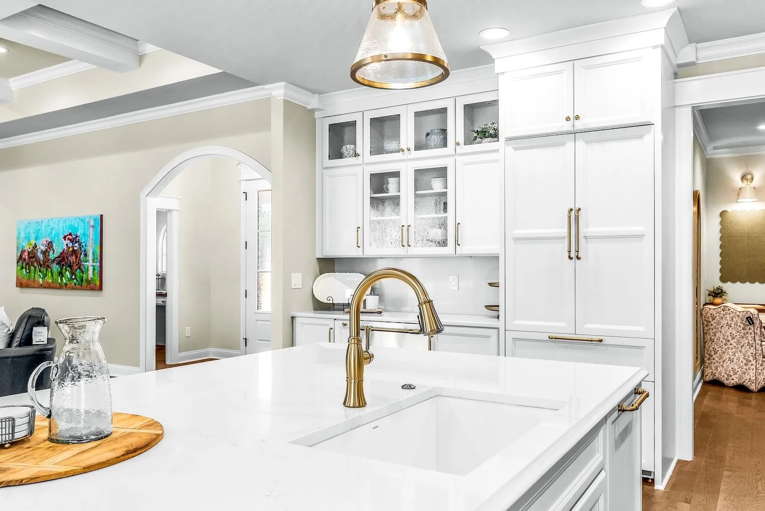Modern kitchen with white cabinetry, gold hardware, and a gold faucet on a white countertop island. Glass-front upper cabinets display dishes. A wooden tray with a glass pitcher and pitcher on the island. Adjacent living space with a colorful horse-r