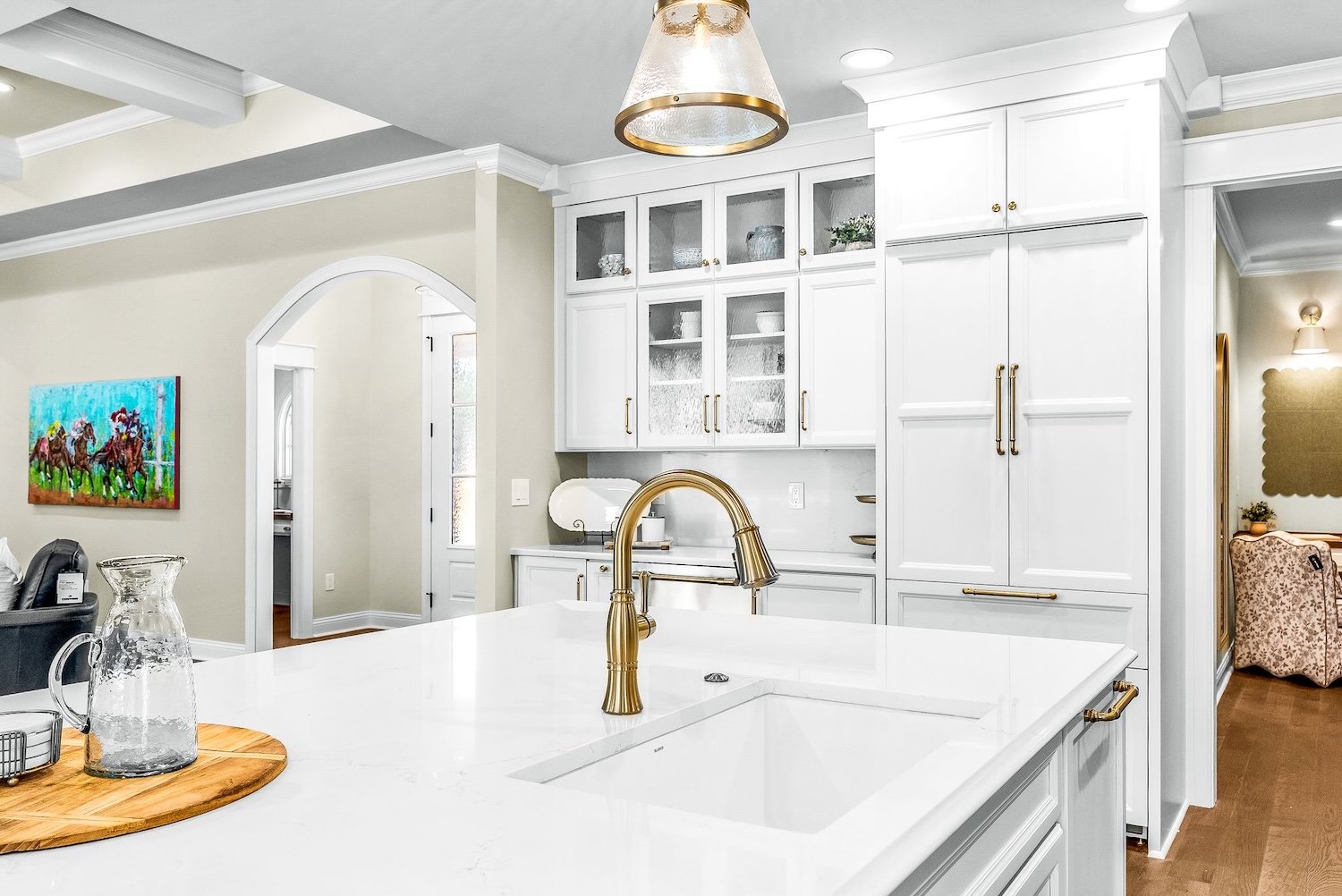 Modern kitchen with white cabinetry, gold hardware, and a gold faucet on a white countertop island. Glass-front upper cabinets display dishes. A wooden tray with a glass pitcher and pitcher on the island. Adjacent living space with a colorful horse-r