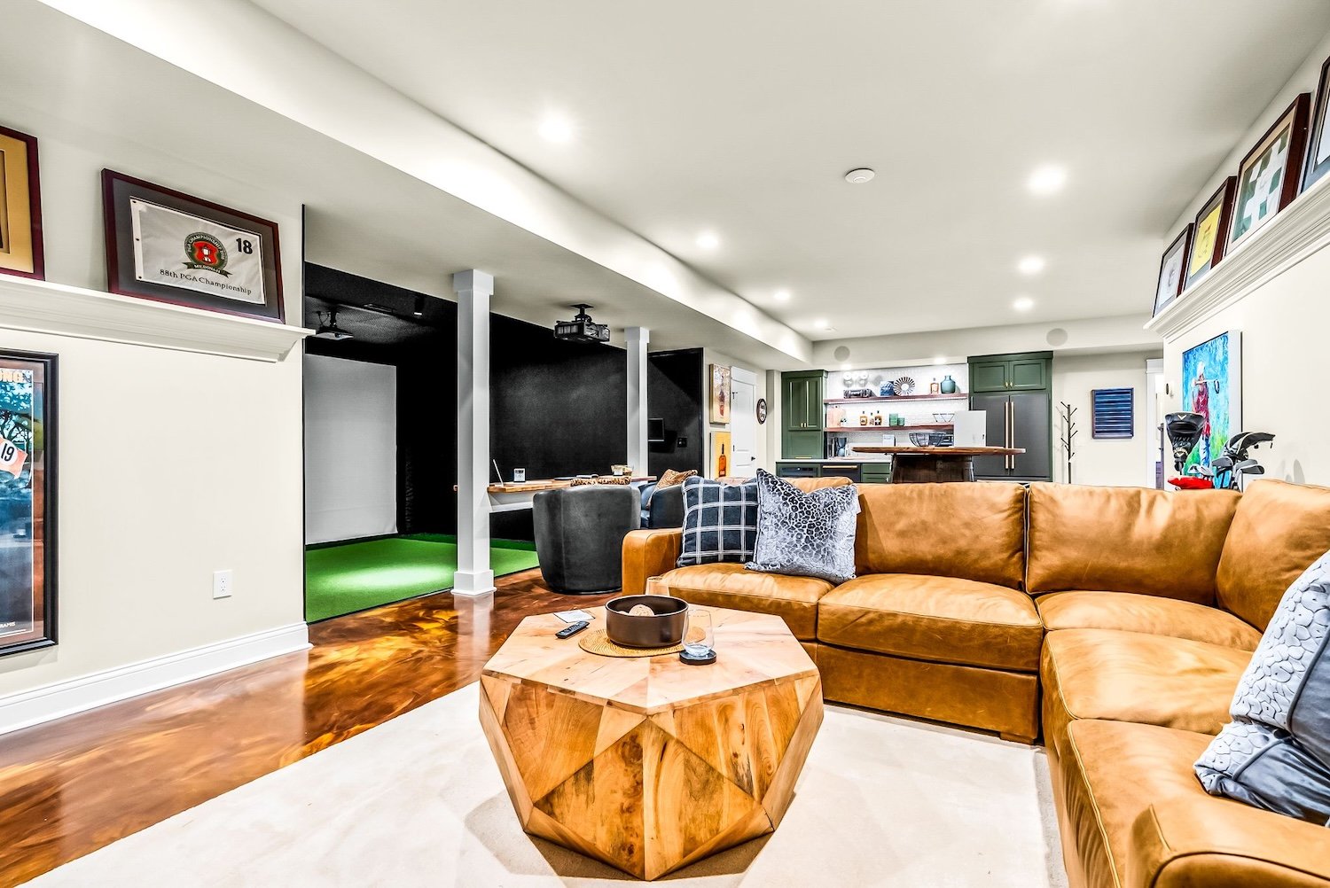 Living room with a tan sectional sofa, wooden geometric coffee table, wall art, and a view of the kitchen with green cabinets and open shelving.
