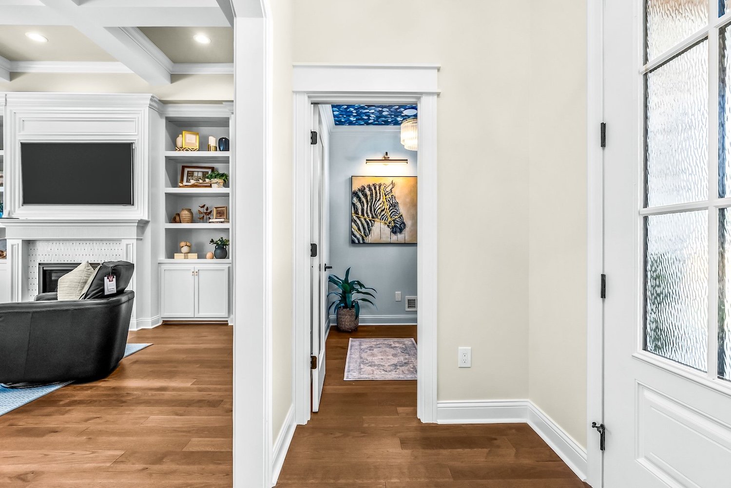 View of a hallway with a doorway leading to a room with a zebra painting, a potted plant, and a small rug. The hallway has light-colored walls and wooden flooring.