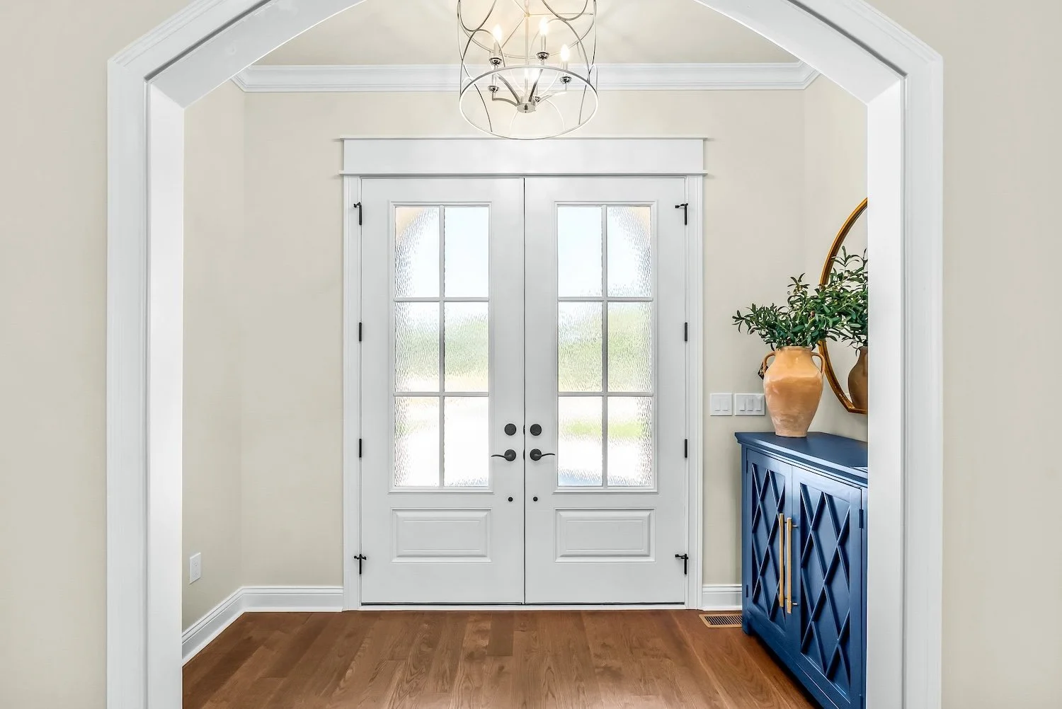Entryway with white double doors, glass panels, beige walls, a dark blue console table with gold handles, a large terracotta vase with green foliage, a round mirror, and hardwood flooring.