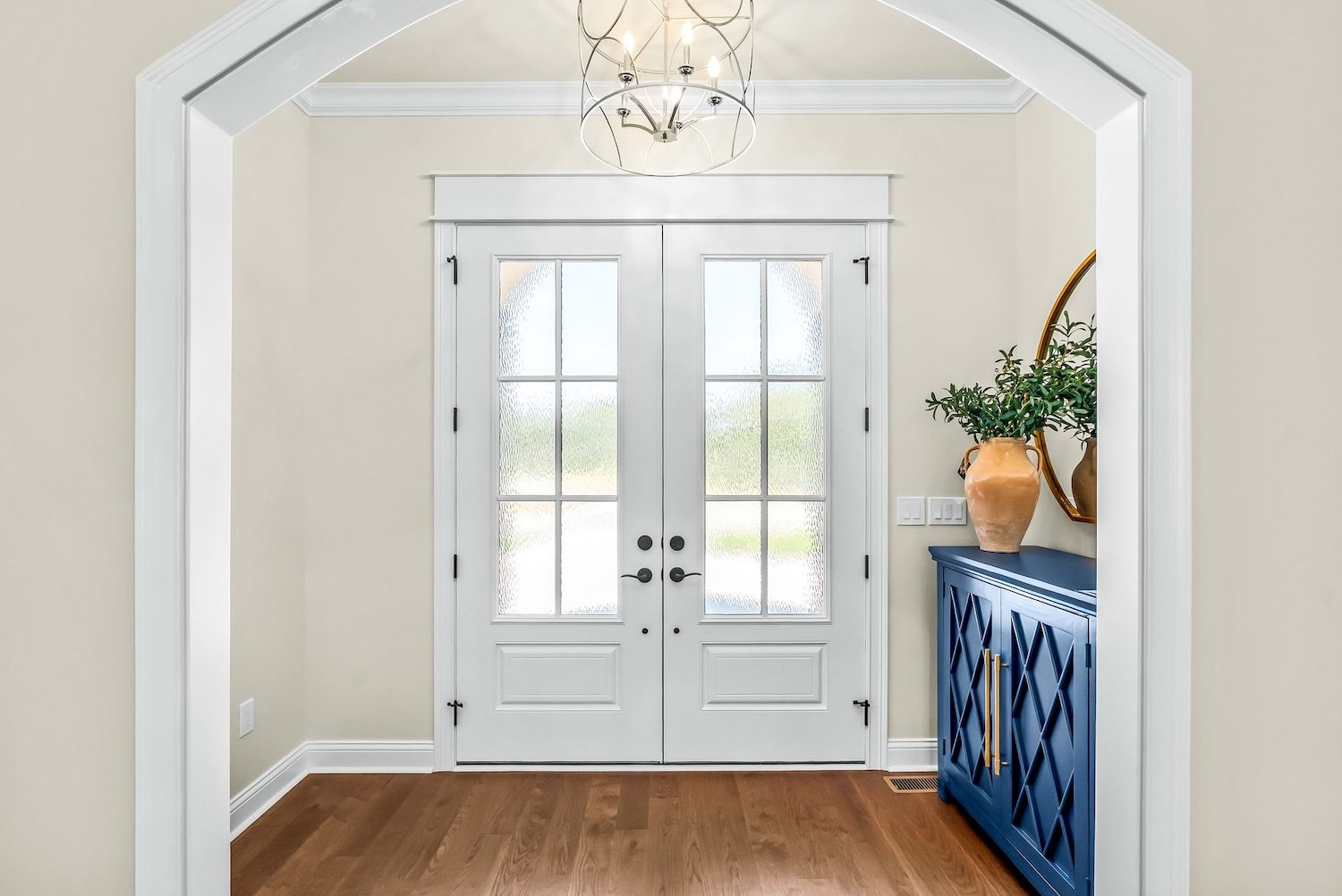 Entryway with white double doors, glass panels, beige walls, a dark blue console table with gold handles, a large terracotta vase with green foliage, a round mirror, and hardwood flooring.