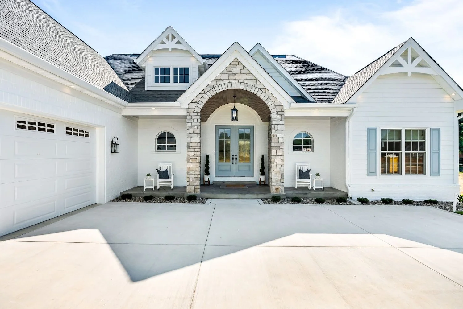 Front view of a modern white house with a stone archway and blue double doors, two benches with cushions, and a landscaped front yard with small bushes.