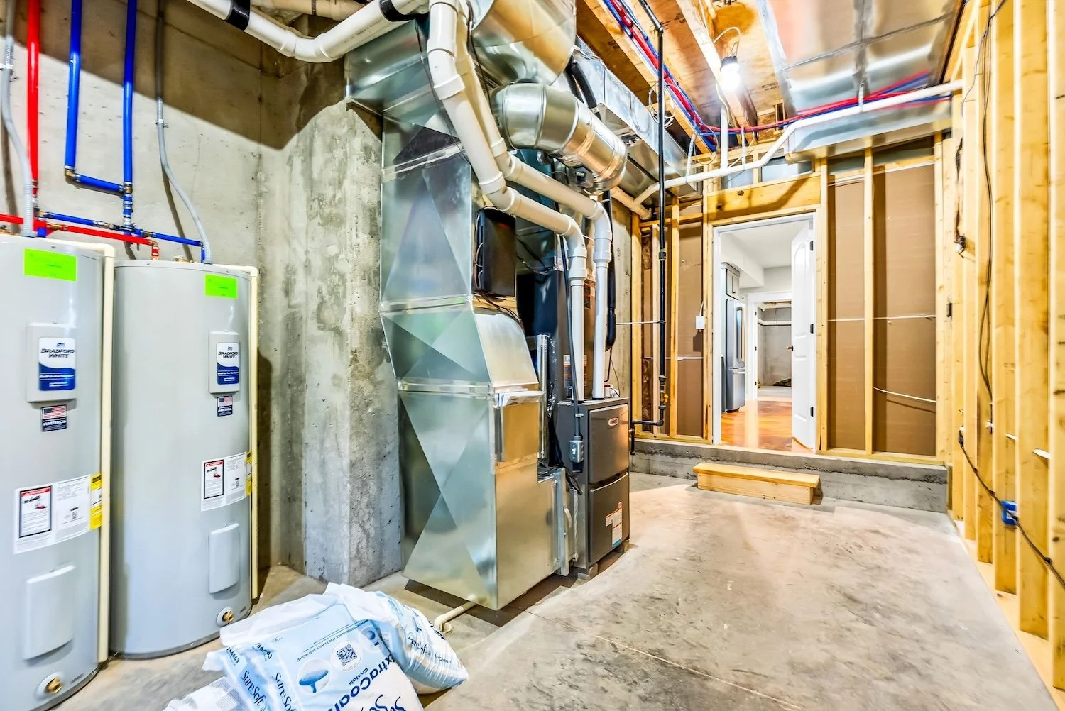 Utility room under construction with water heaters, ductwork, and framing. Doorway to a room with hardwood floors.