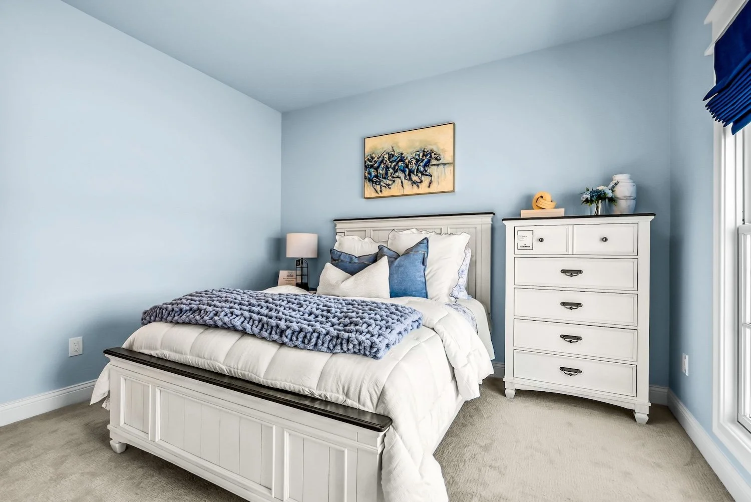A bedroom with light blue walls, white wooden bed frame, and a dresser. The bed has white bedding, multiple pillows in shades of blue and white, and a chunky knit gray blanket. A nightstand with a lamp is beside the bed, and a painting of horse racin