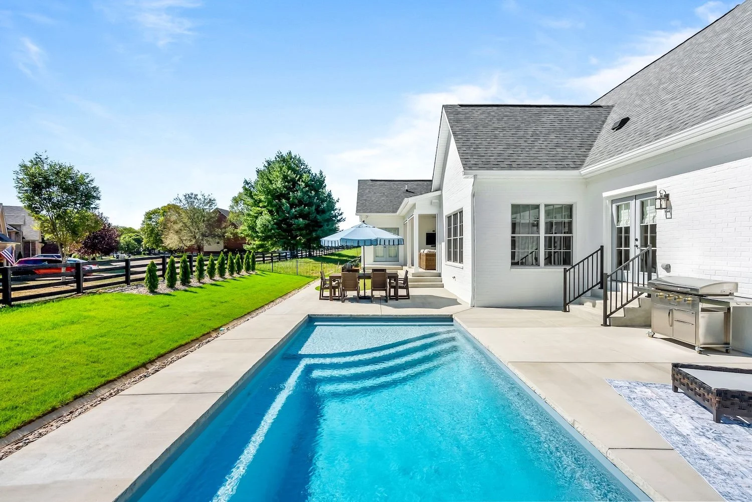 Backyard with a swimming pool, patio furniture with a large blue umbrella, green lawn, trees, white house with stairs leading to a door, and clear blue sky.