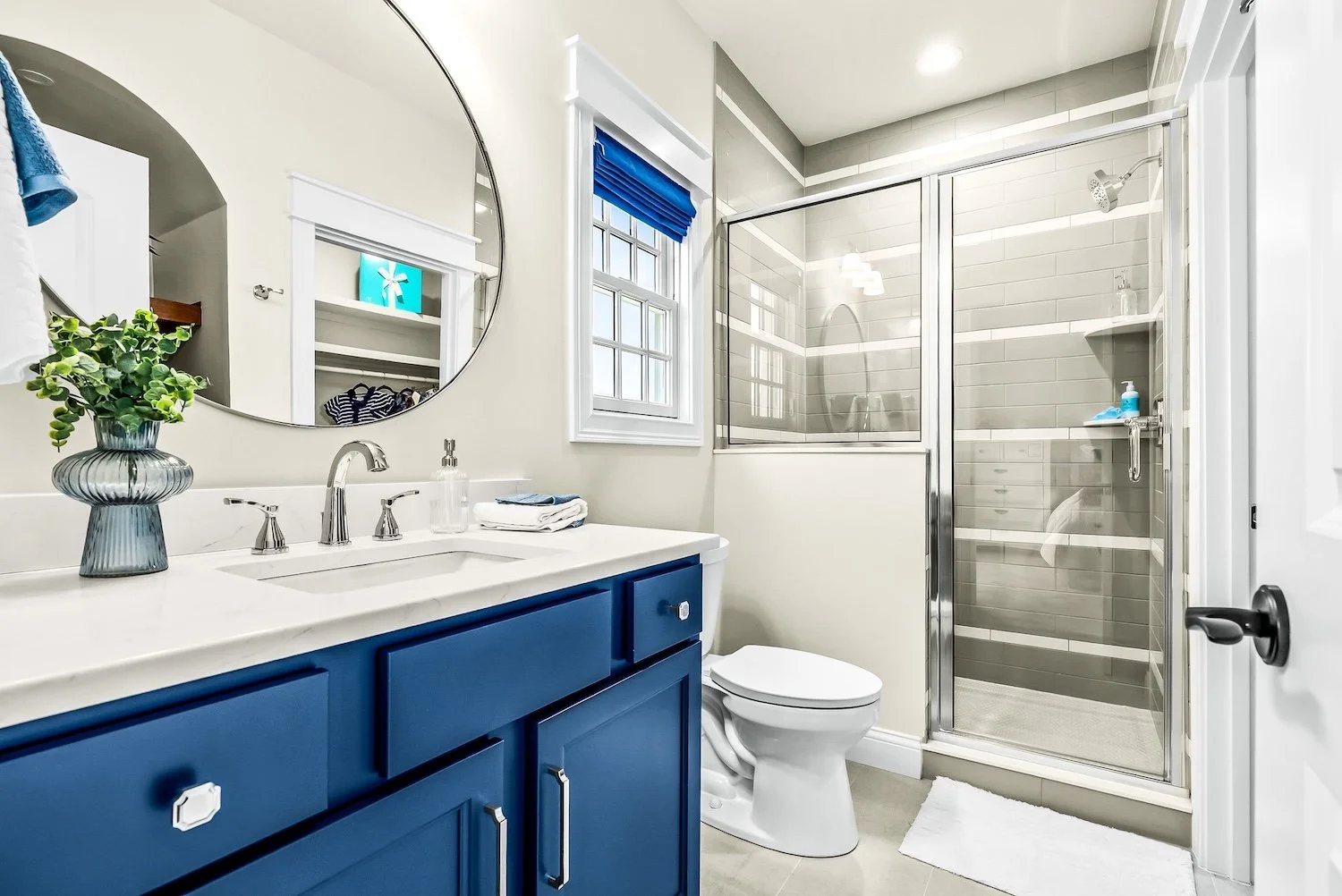 Bathroom with a blue cabinet vanity, oval mirror, white countertop, toilet, and a glass-enclosed shower with gray tiles.