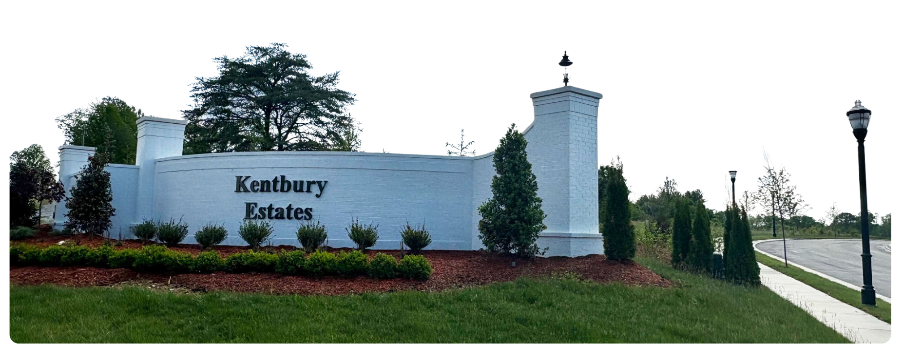 Entrance sign for Kentbury Estates with white brick wall, black lettering, and landscaped greenery including shrubs, trees, and grass, along with lamp posts along the sidewalk.
