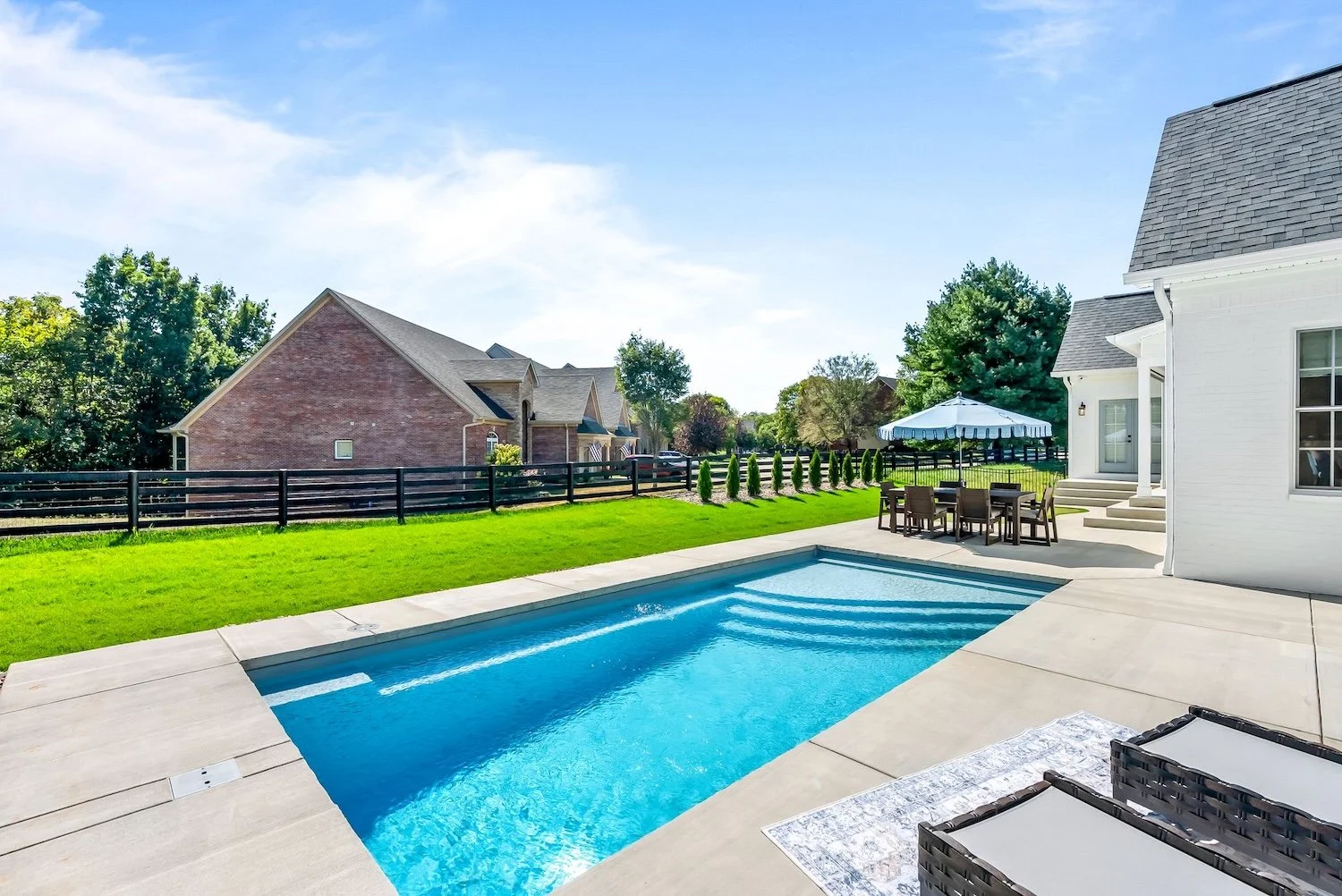 Residential backyard with a swimming pool, patio furniture, a striped umbrella, a lawn, and neighboring houses with trees under a blue sky.