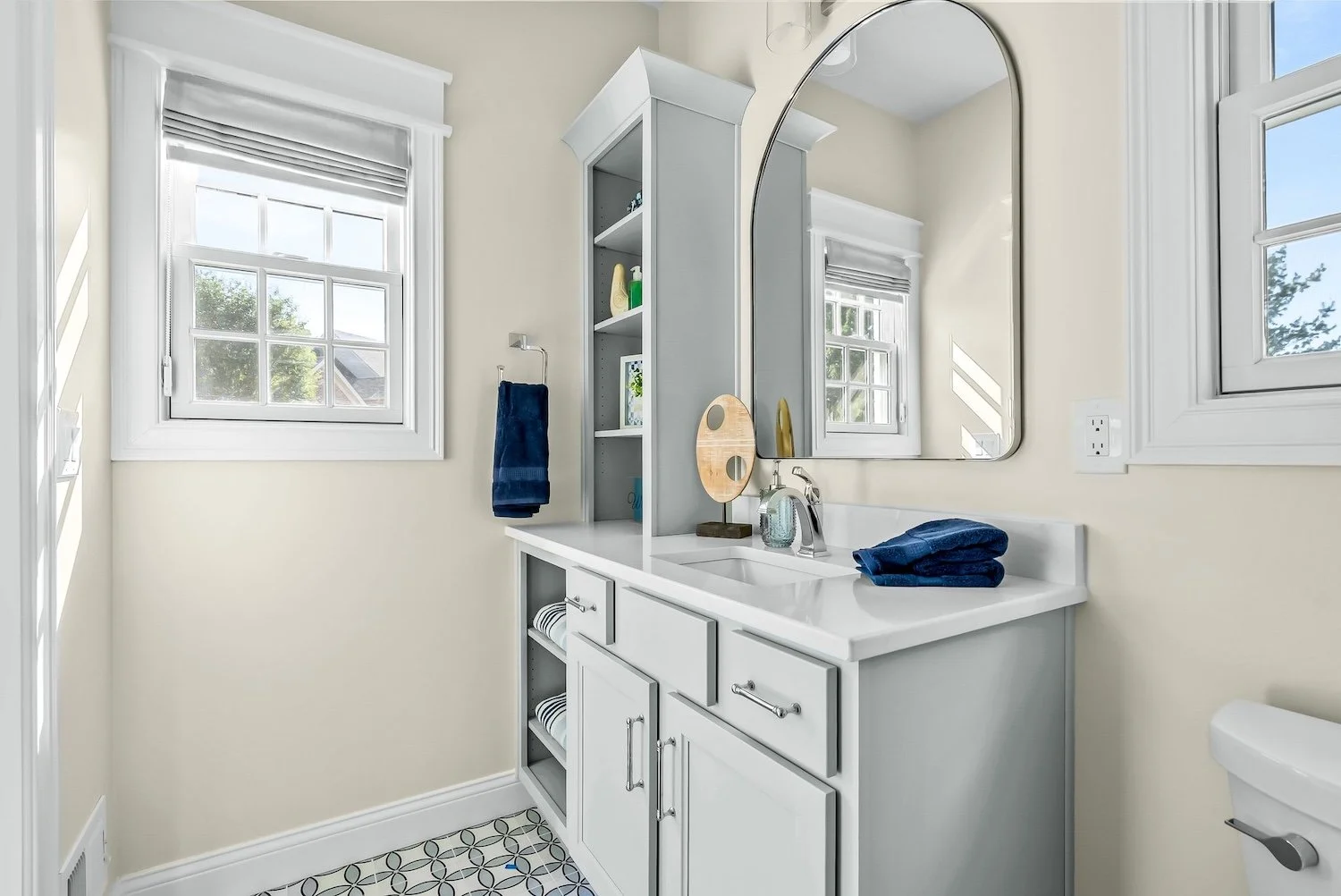 Bright bathroom with white vanity, oval mirror, and two windows with white trim and blinds. Blue towels on the counter, towel hanging on wall, patterned floor tiles, and shelves with towels and toiletries.