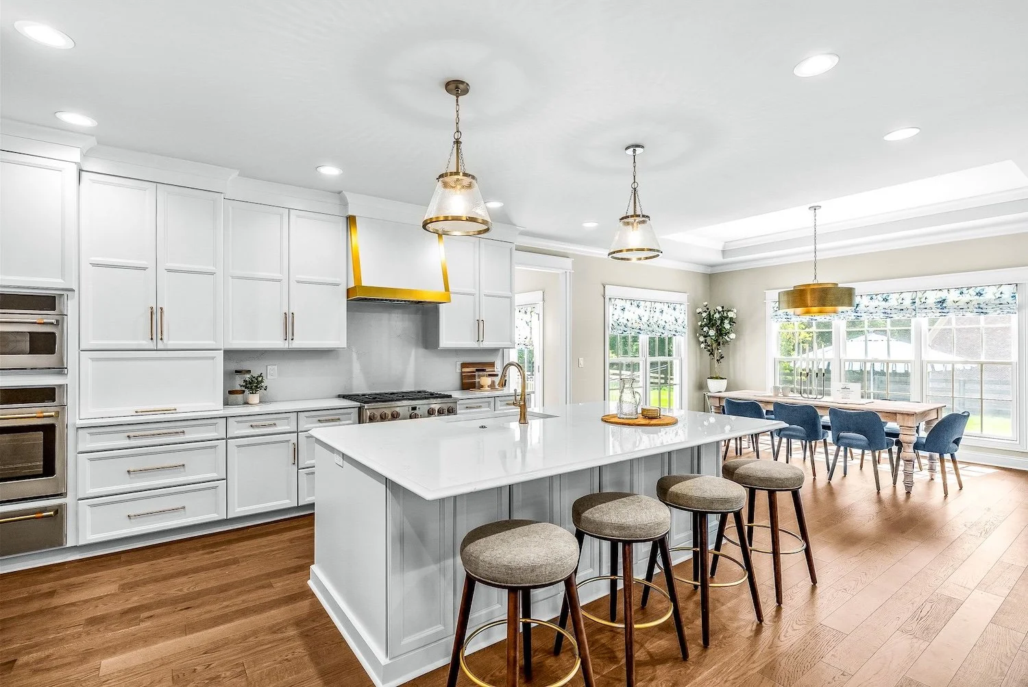 Modern kitchen with white cabinets, a large island with a white marble countertop, wooden stools, and a dining area with a wooden table and blue chairs, surrounded by large windows and natural light.