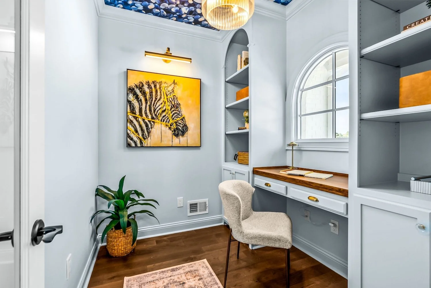 Small home office with a window, built-in shelves, a desk with a wooden top, a beige upholstered chair, a potted plant, a framed zebra artwork, and a ceiling light fixture.