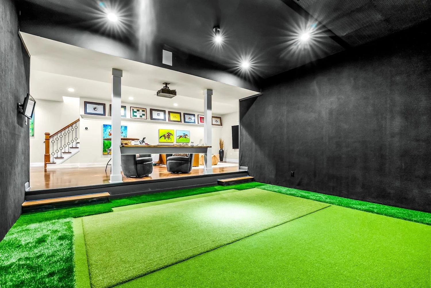 An indoor golf simulator room with green artificial turf, black walls, and ceiling. The room opens to a bright living area with artwork, a staircase, and two black chairs.