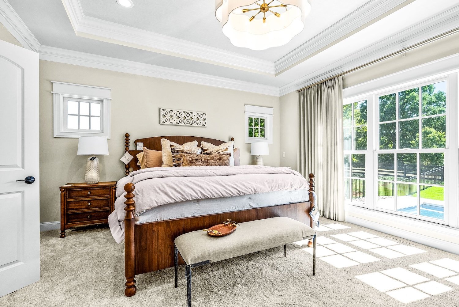 Bright bedroom with large window view of green yard, wooden bed with beige bedding, tufted bench at foot of bed, two nightstands with lamps, light-colored walls, ceiling with decorative molding and chandelier.
