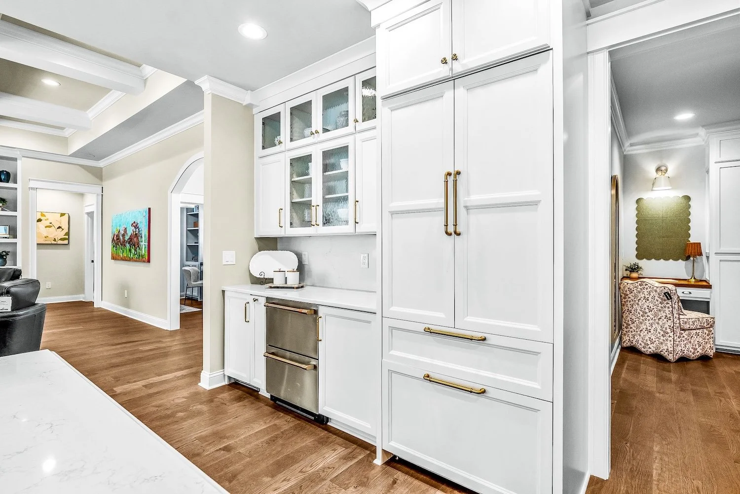 Modern kitchen with white cabinets, gold handles, and hardwood floors, leading to a cozy sitting area with a floral armchair and a wall mirror.