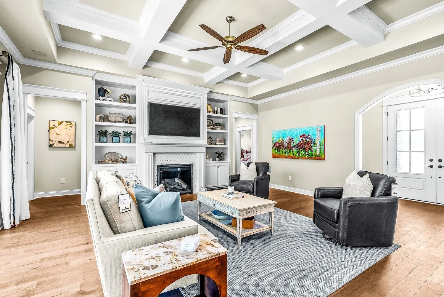 Living room with beige walls, white ceiling with coffered design, wooden ceiling fan, built-in white shelves, a fireplace, a flat-screen TV, a beige sofa with pillows, two black armchairs, a coffee table, a rug, and wall art depicting horses.
