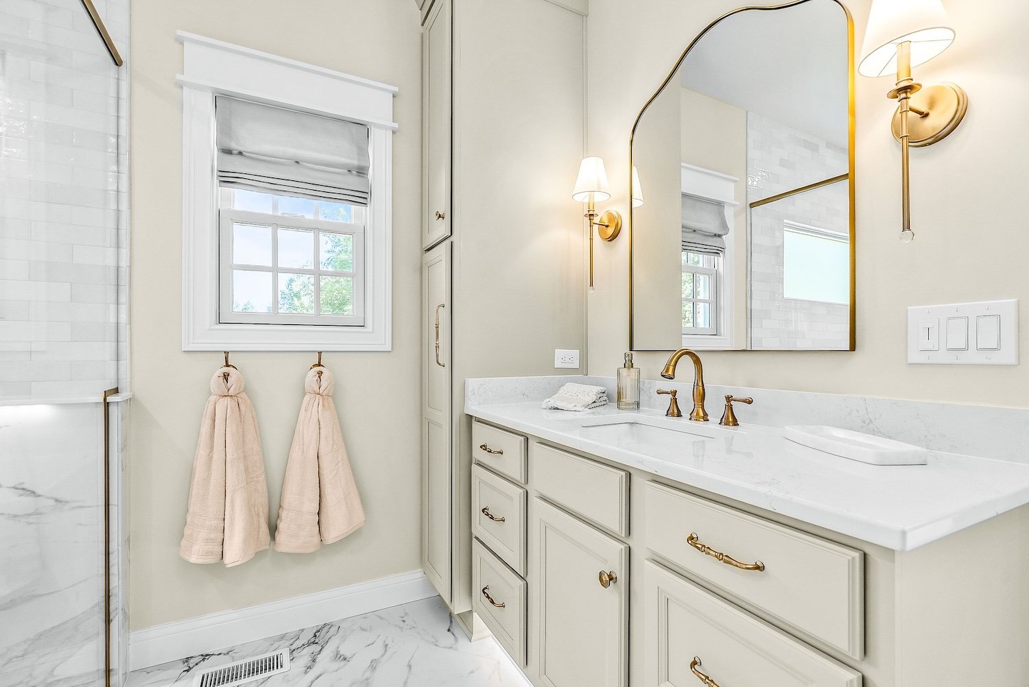Bathroom with a white vanity, gold fixtures, a large mirror, wall-mounted lights, a window with a Roman shade, beige towels, and marble flooring.