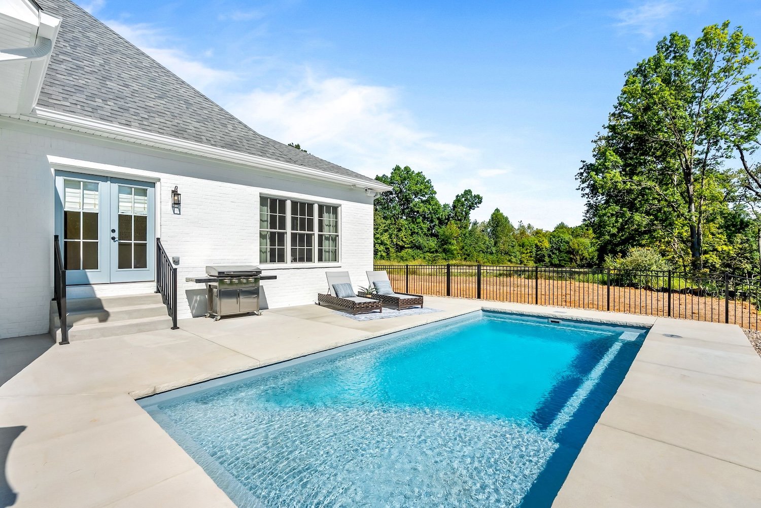 Residential backyard with a white house, gray roof, swimming pool, two lounge chairs, a grill, and a fenced backyard with trees and open sky.