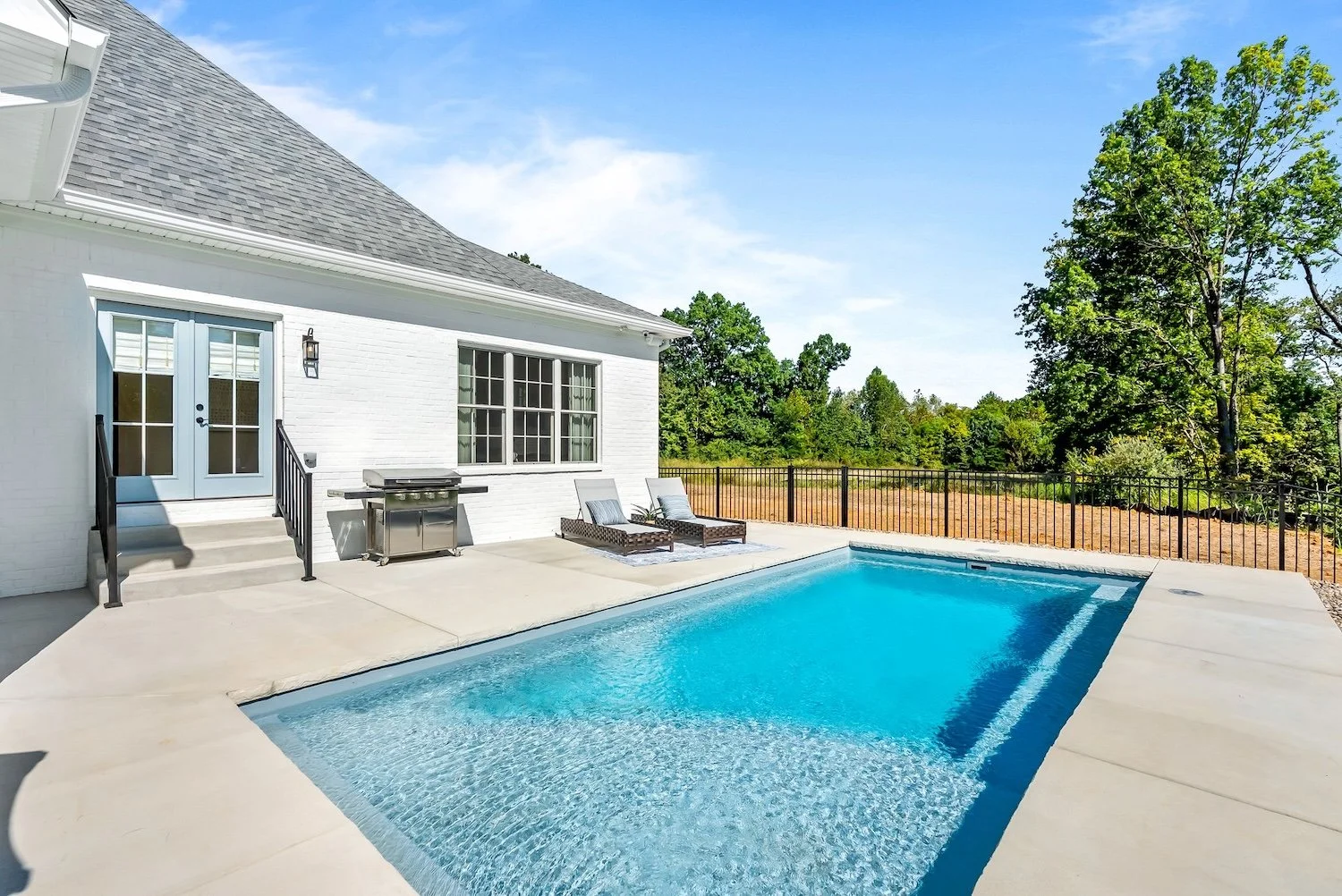 Residential backyard with a white house, gray roof, swimming pool, two lounge chairs, a grill, and a fenced backyard with trees and open sky.
