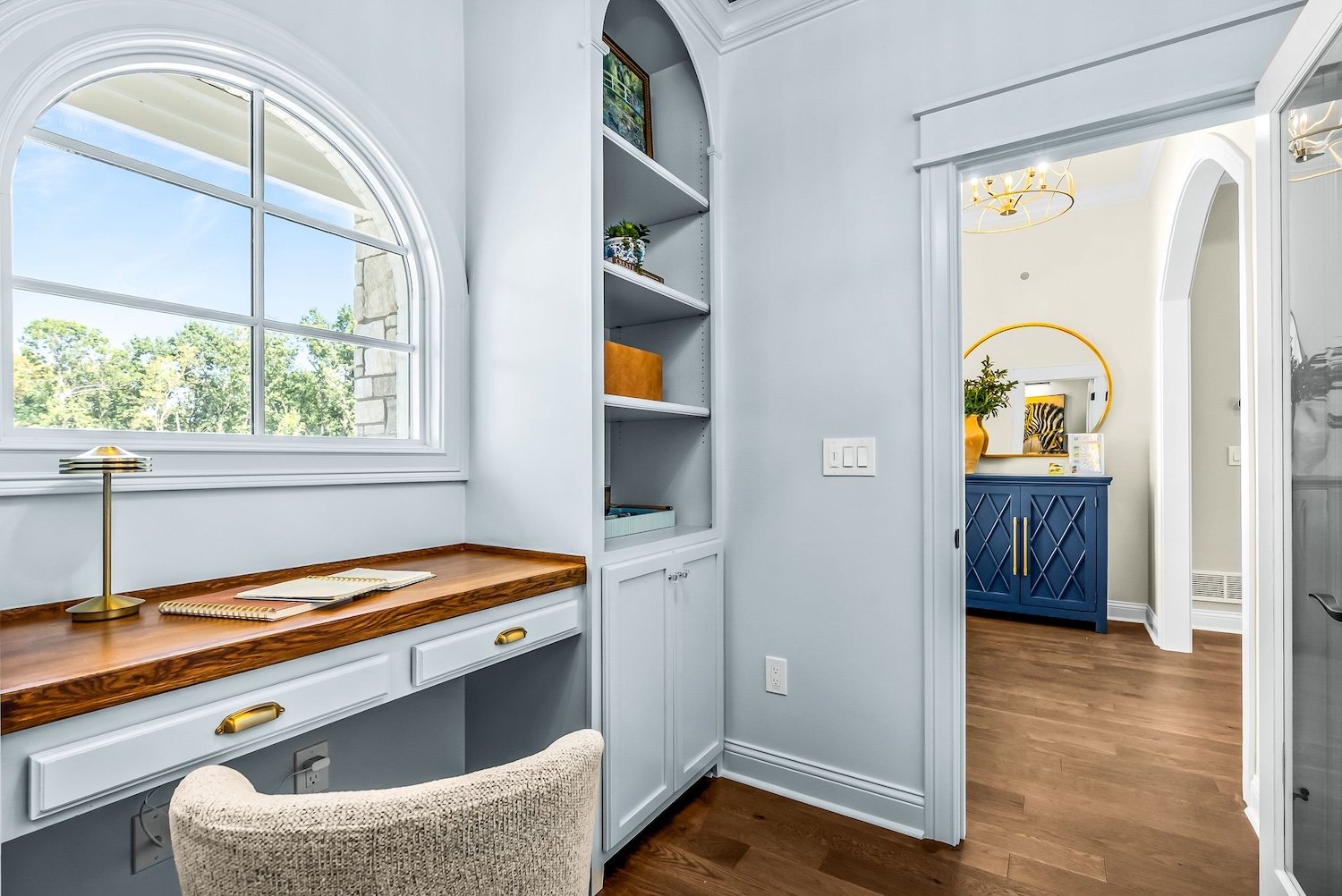 Home interior featuring a window with decorative molding, a wooden desk with a lamp and notebooks, built-in shelves, and a view into another room with a blue sideboard and a round mirror.