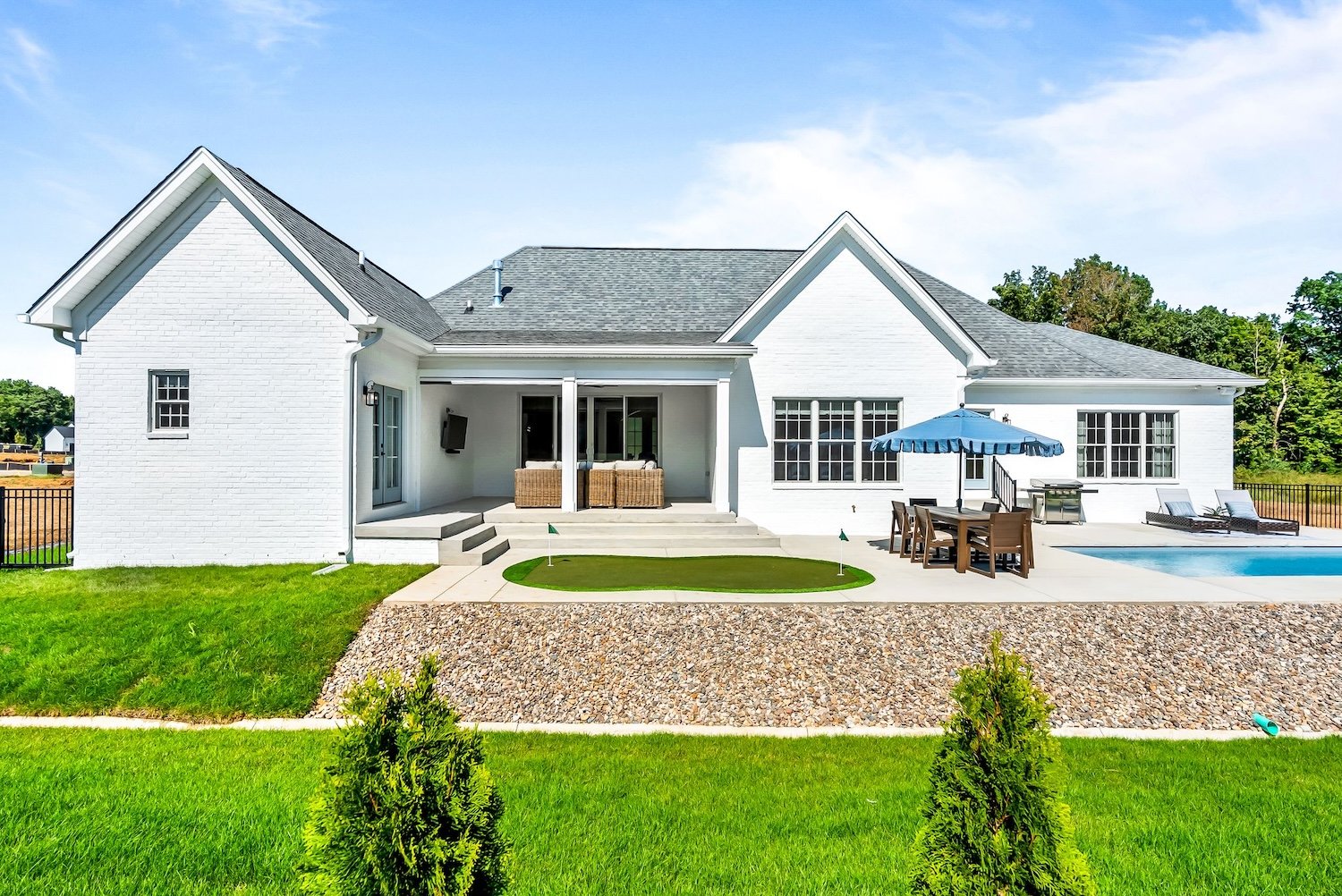 Front view of a white brick house with a backyard patio, outdoor furniture, a swimming pool, green lawn, and small trees.