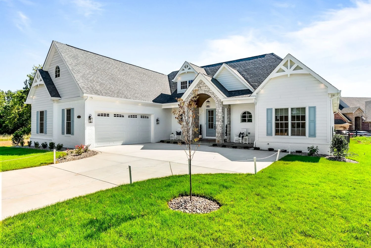 Front view of a white, modern house with a gray roof, front porch, two chairs, and a small tree in the front yard, on a sunny day with green grass.