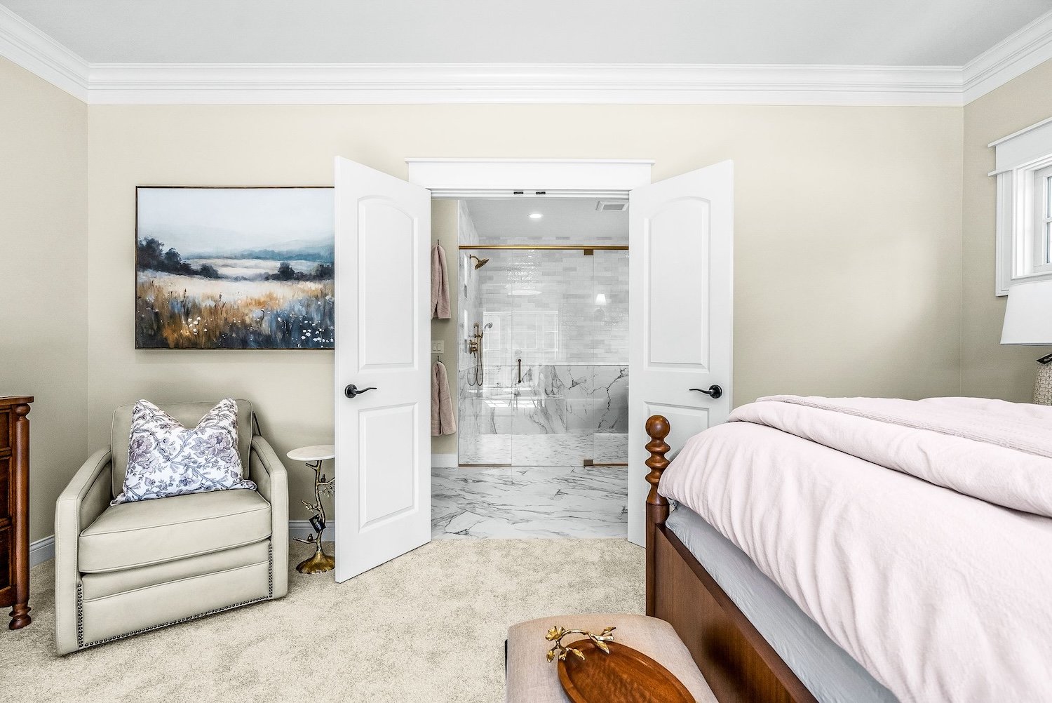 A bedroom with a large bed, white bedding, a wooden bedside table, and a cream armchair with a pillow, with an open door leading to a marble-floored bathroom.