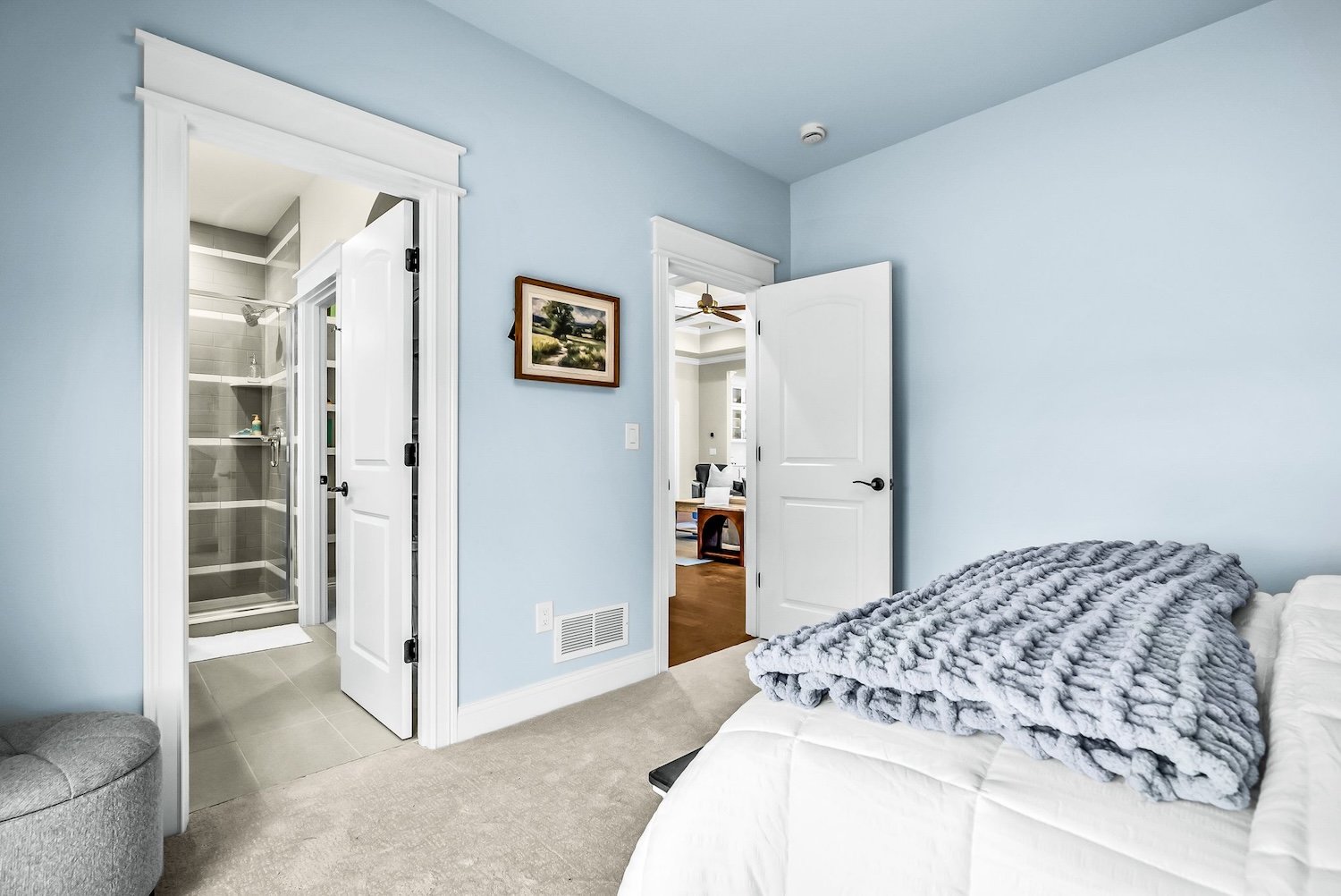 A bedroom with light blue walls, a bed with white sheets and a gray faux fur blanket, an open door leading to a hallway, a bathroom with a shower visible, a framed landscape painting on the wall, and a beige carpeted floor.