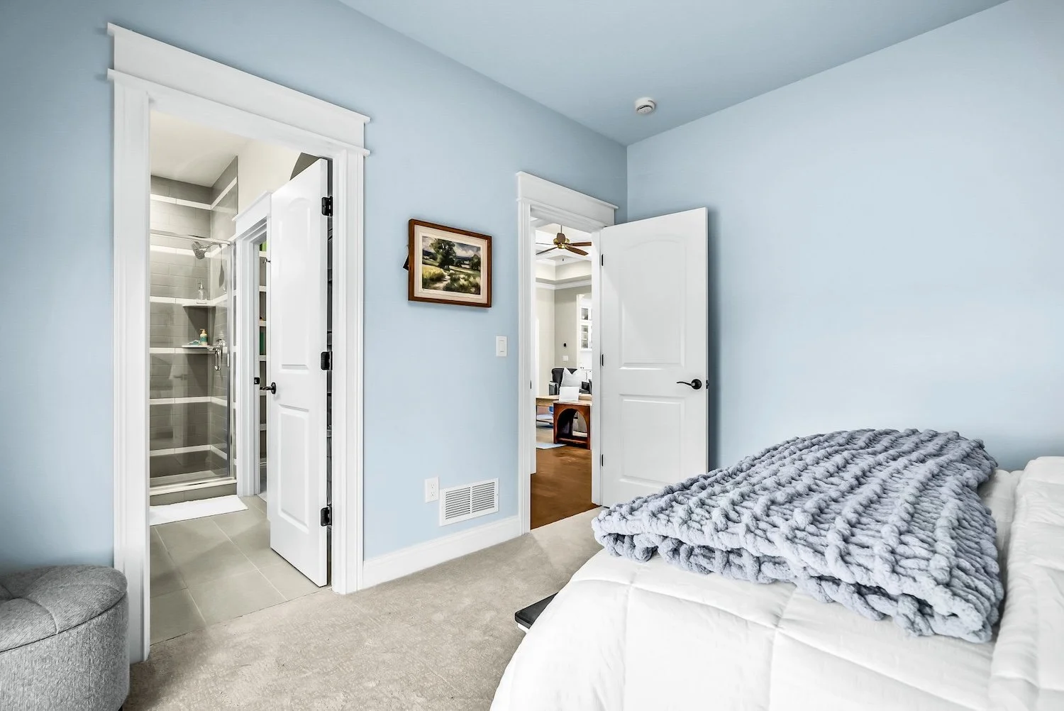 A bedroom with light blue walls, a bed with white sheets and a gray faux fur blanket, an open door leading to a hallway, a bathroom with a shower visible, a framed landscape painting on the wall, and a beige carpeted floor.