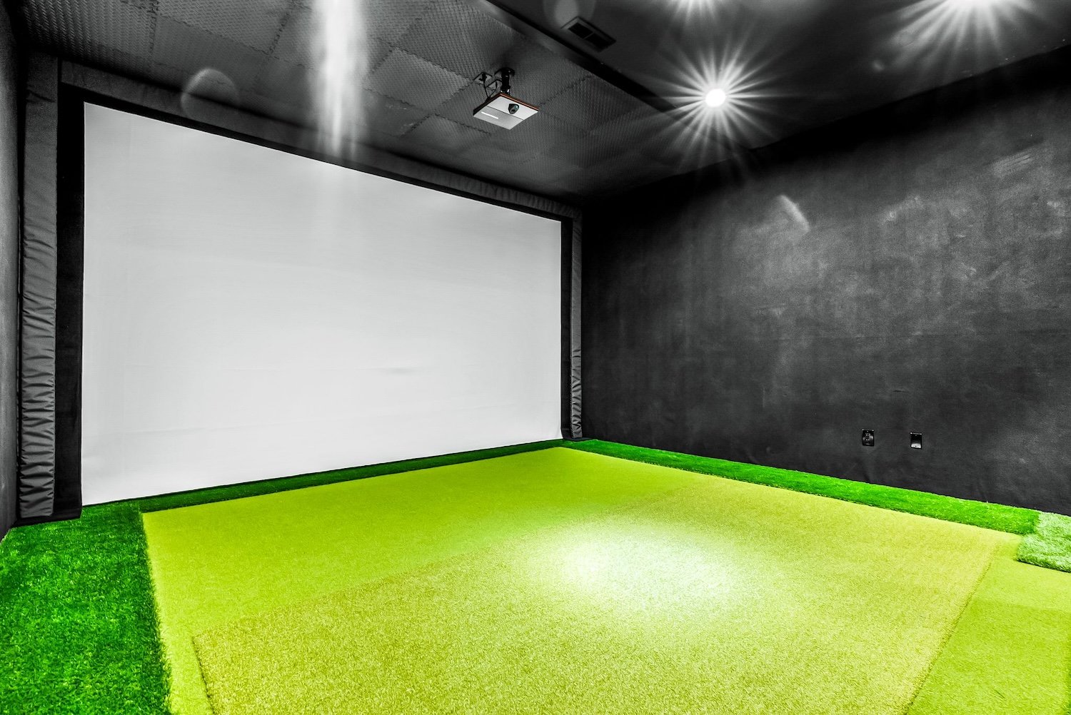 A black and white movie theater room with a large white projection screen, a ceiling-mounted projector, and green artificial turf on the floor.