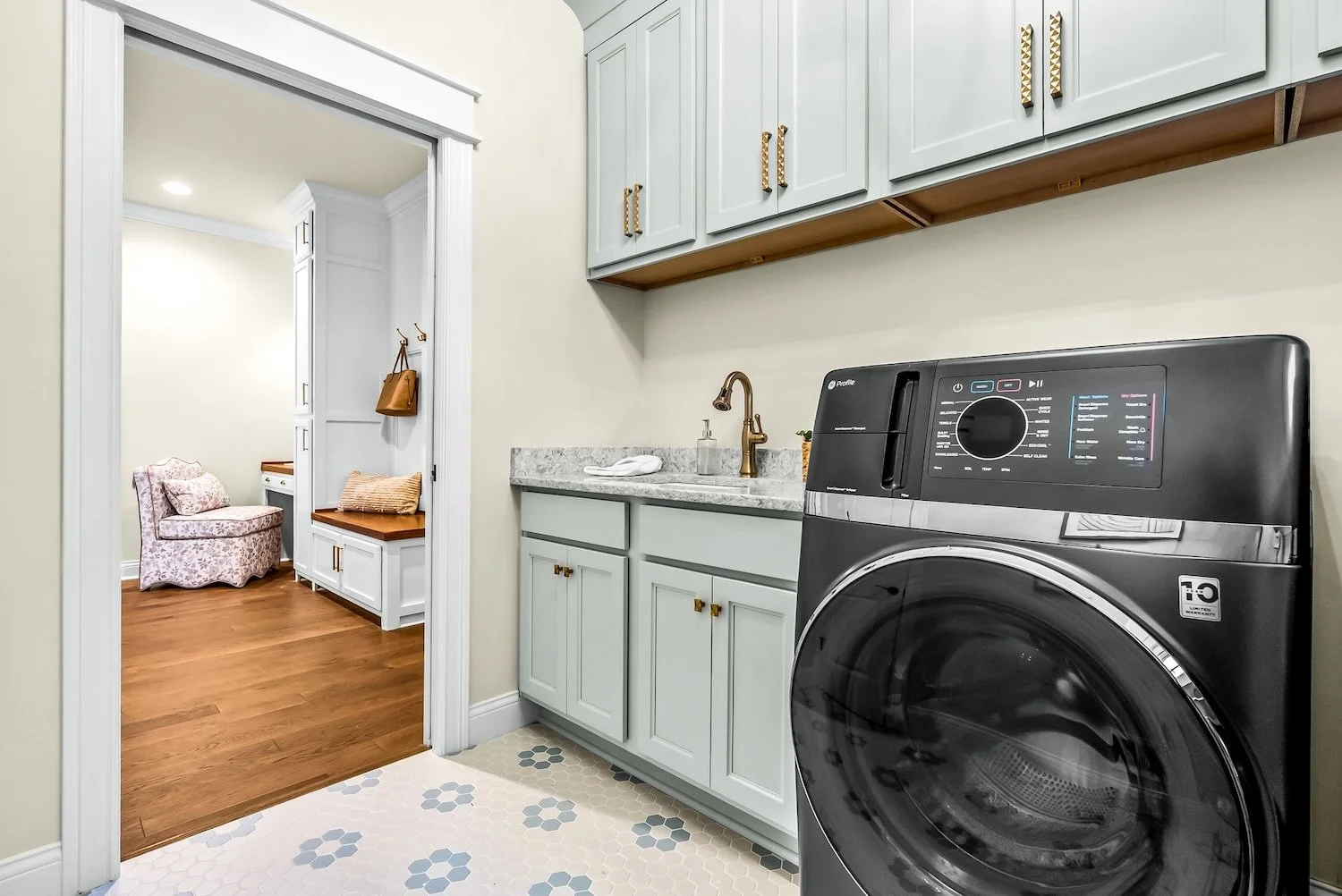 Laundry room with front-loading washing machine, white cabinets, marble countertop, brass faucet, and a doorway leading to a sitting area with an armchair and built-in bench.