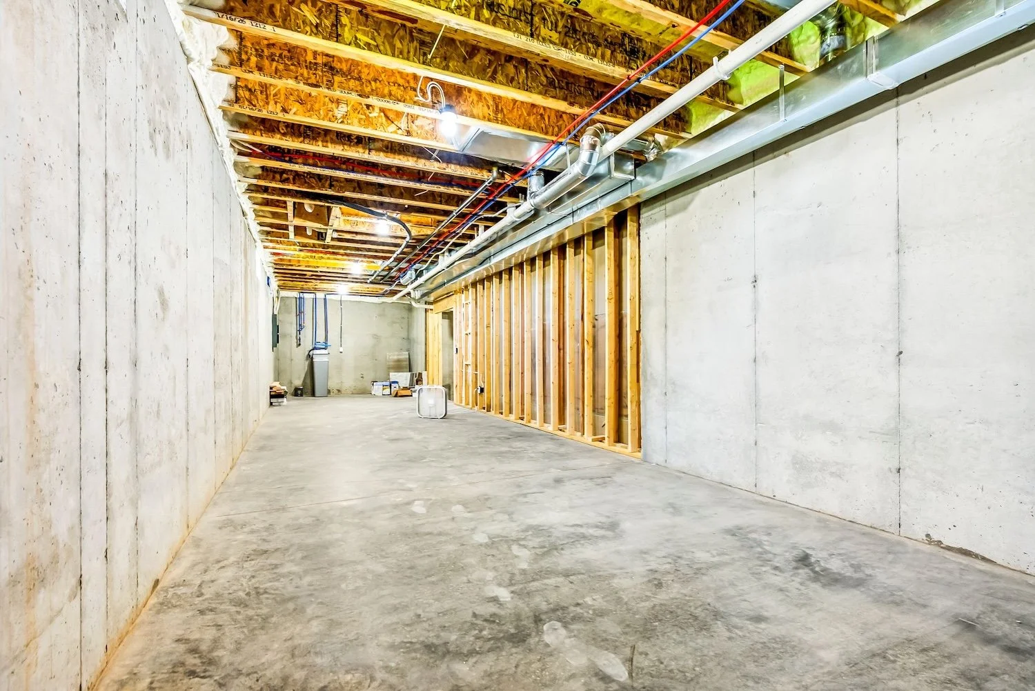 Unfinished basement with concrete walls, exposed ceiling with wooden beams and pipes, empty concrete floor, and some construction materials in the background.
