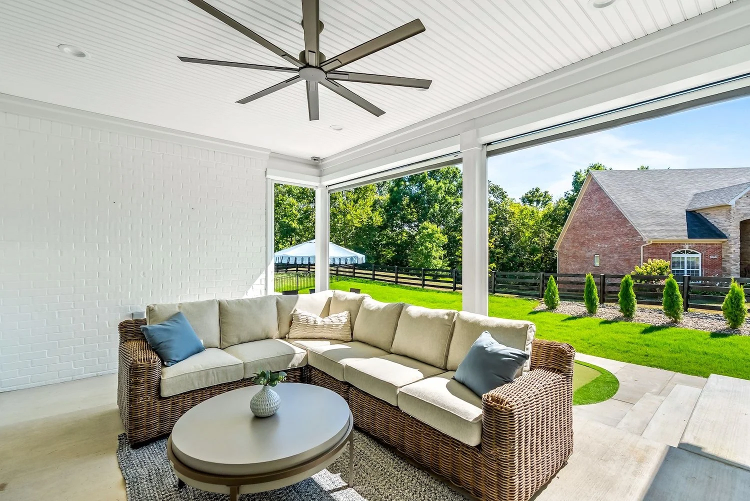 A screened-in porch with a white brick wall, beige sectional couch with blue and beige throw pillows, a round coffee table with a small potted plant, ceiling fan, and a view of a green backyard with trees, shrubs, a black fence, and neighboring house