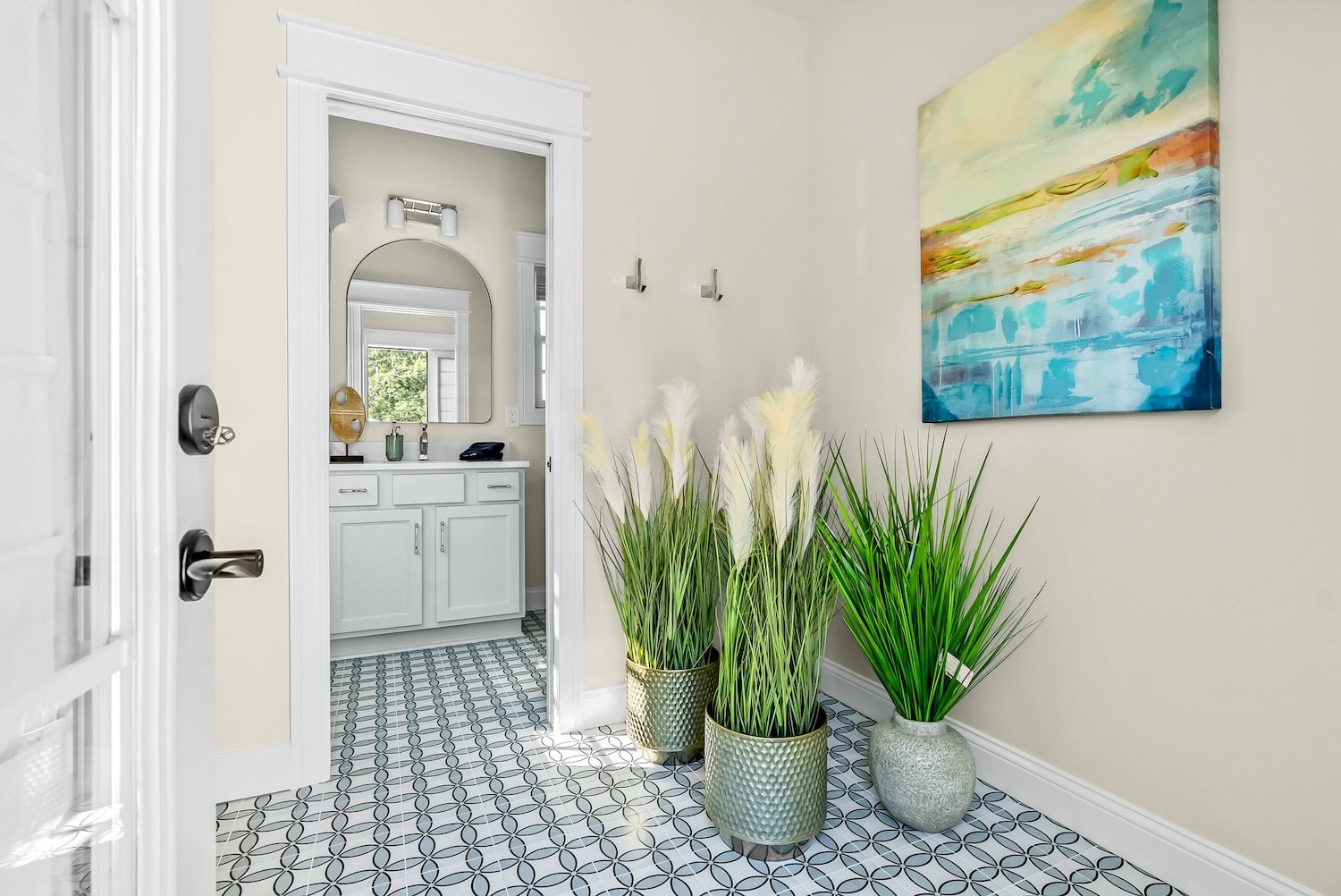 Entryway with three large potted plants, off-white walls, a multi-colored abstract painting on the wall, and a view into a bathroom with white cabinetry and geometric patterned floor tiles.