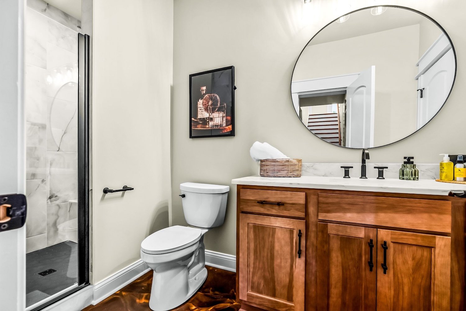 A modern bathroom with a white toilet next to a wooden vanity with a marble countertop, a round mirror, and a sail bottle, black fixtures, and framed artwork on the wall.