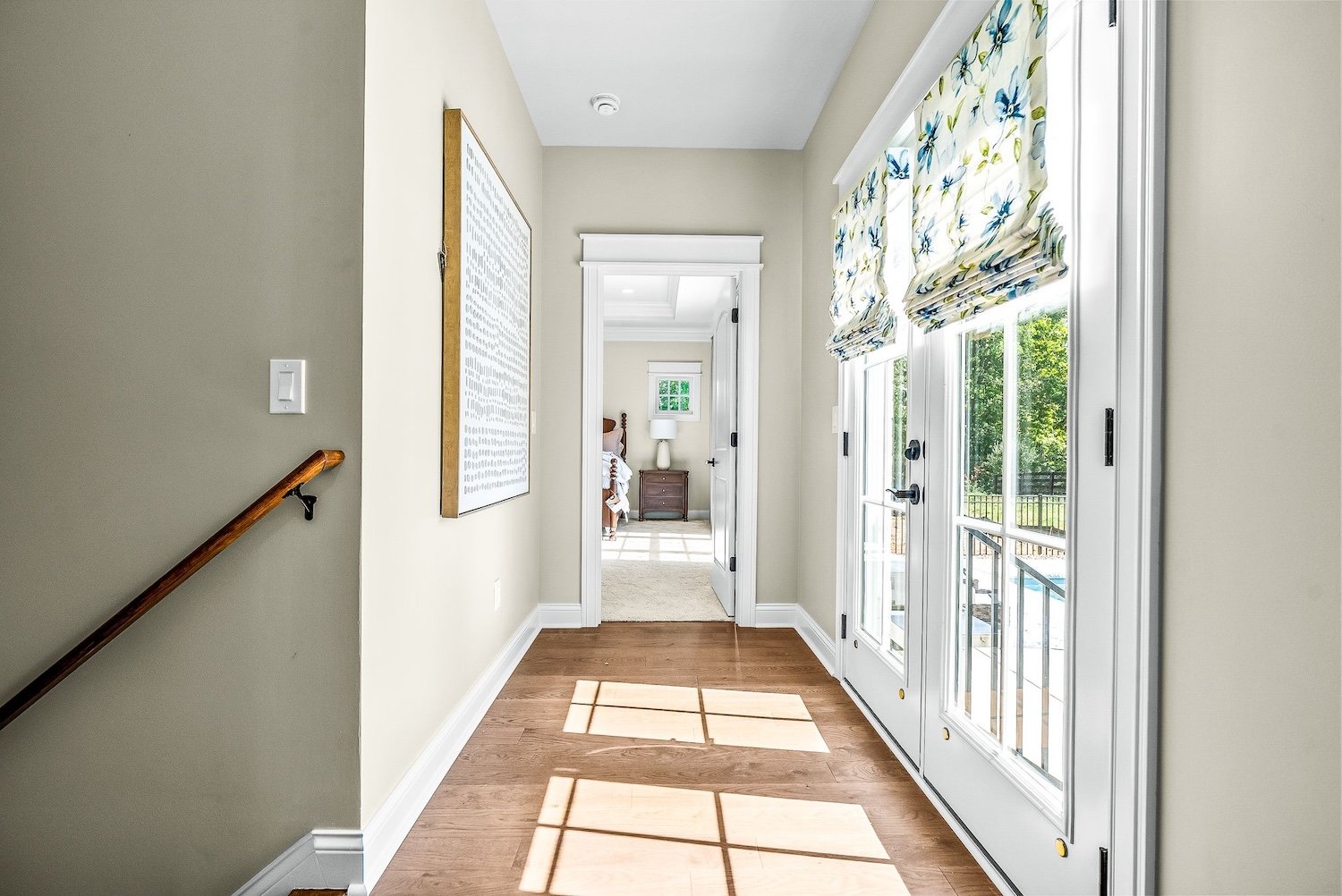 Bright hallway with wooden flooring, French doors with floral curtains leading outside, and a bedroom at the end with a wooden nightstand and lamp.