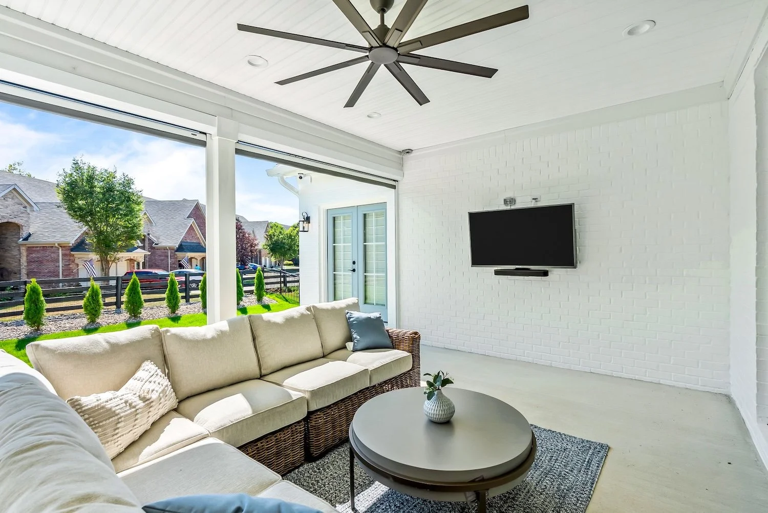 Sunlit enclosed porch with white brick wall, wall-mounted TV, ceiling fan, beige sofa with pillows, round coffee table with plant, view of neighborhood with houses and trees outside.