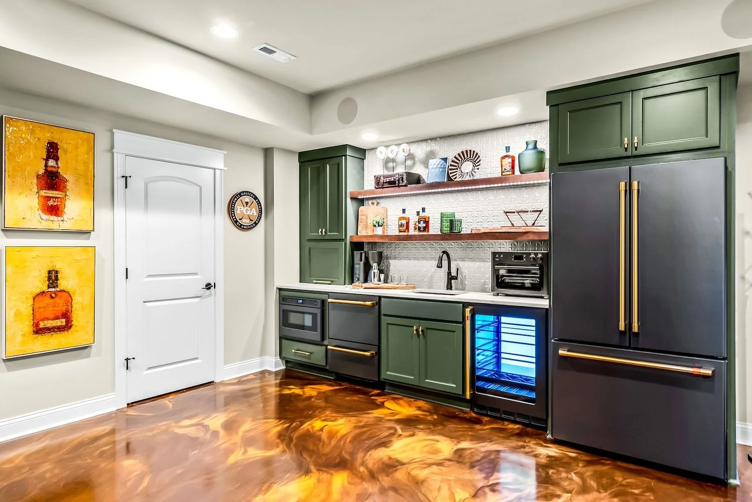 Kitchen with green cabinets, black refrigerator with gold handles, small wine fridge, white backsplash tiles, open shelving with decor and bottles, artwork on walls, hardwood floor with swirling wood grain pattern.