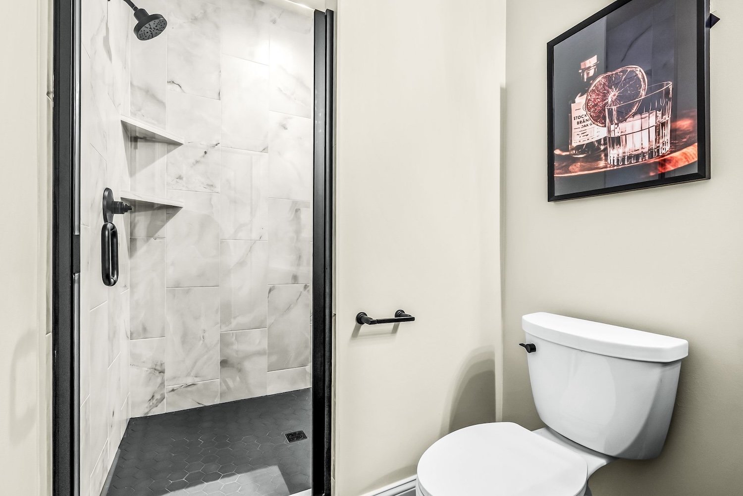 Bathroom with a shower area that has gray and white marble tiles, a black showerhead, a black shower handle, and a small built-in shelf. Next to the shower is a white toilet and a framed picture of a glass of whiskey with a slice of citrus on the wal
