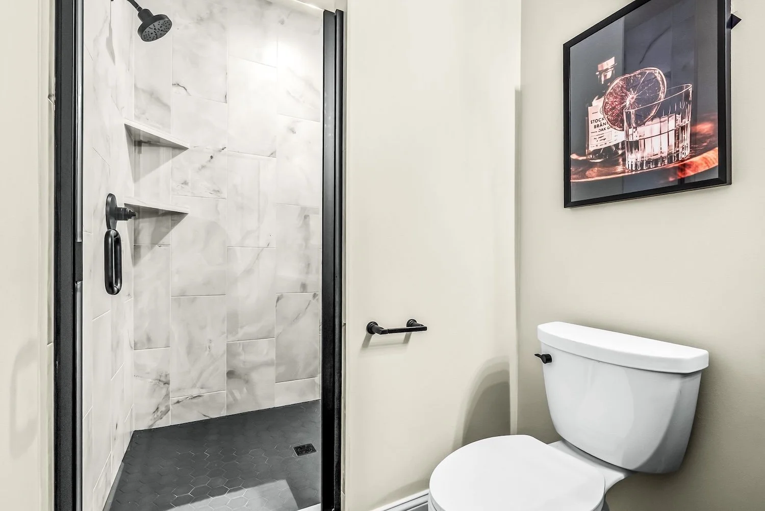 Bathroom with a shower area that has gray and white marble tiles, a black showerhead, a black shower handle, and a small built-in shelf. Next to the shower is a white toilet and a framed picture of a glass of whiskey with a slice of citrus on the wal