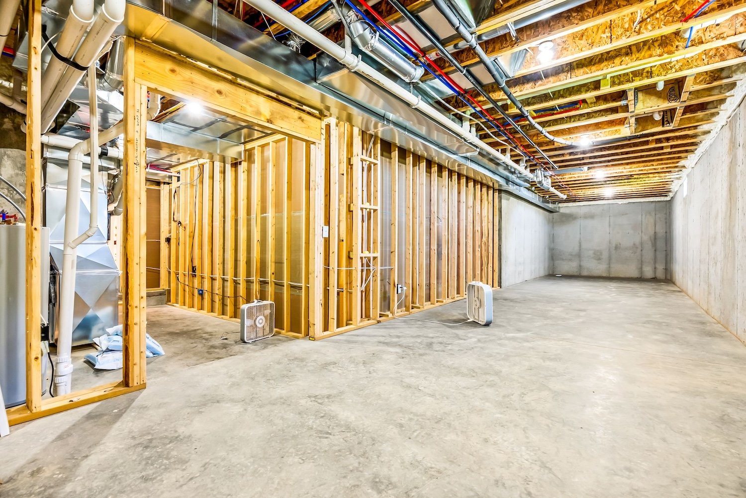 An unfinished basement with exposed wooden framing, concrete walls, and ceiling joists. There are two small fans on the floor, electrical wiring, and plumbing pipes, with some insulation visible. The space is empty and under construction.