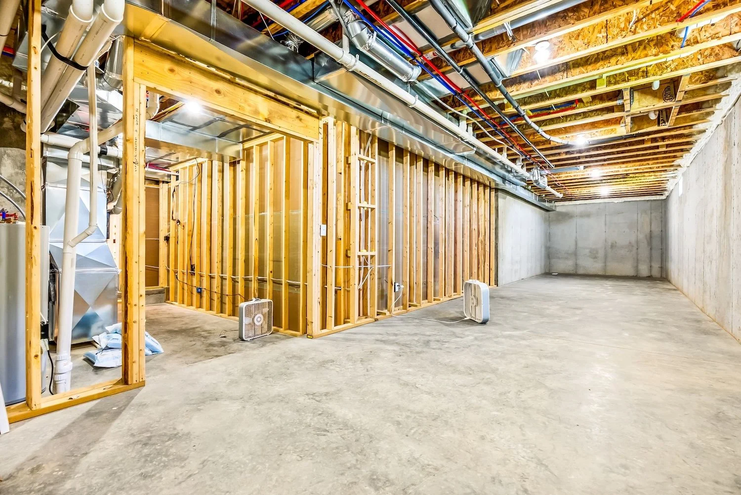 An unfinished basement with exposed wooden framing, concrete walls, and ceiling joists. There are two small fans on the floor, electrical wiring, and plumbing pipes, with some insulation visible. The space is empty and under construction.