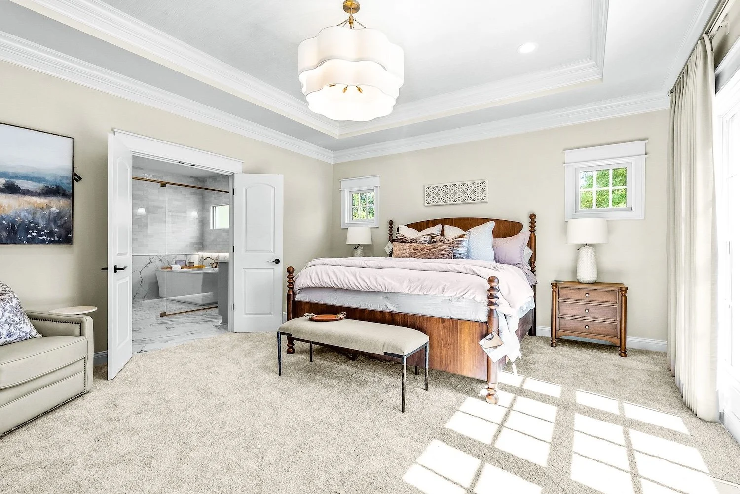 A cozy bedroom with a large wooden bed, white bedding, and multiple pillows, illuminated by natural light through a sliding glass door and windows, with a small bench at the foot of the bed, a nightstand with a lamp, and an open door leading to a bat