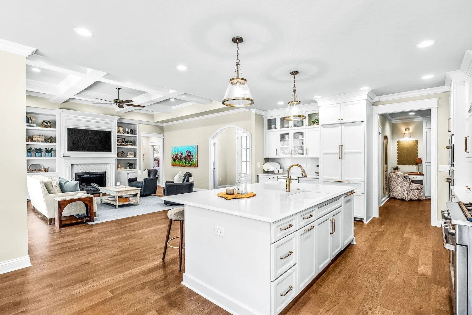 Open-concept kitchen and living room with white cabinetry, hardwood floors, central island, pendant lighting, and a cozy seating area with a fireplace and television.